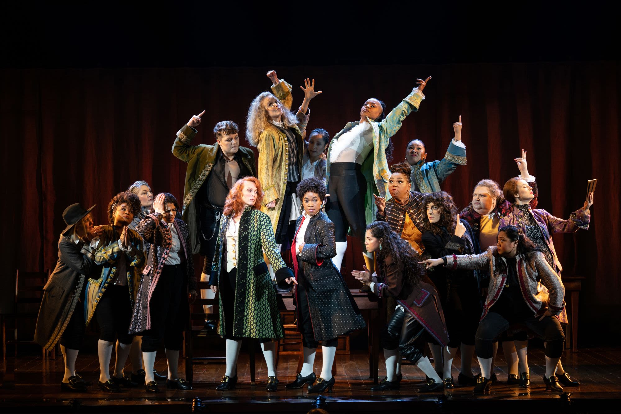 The company of Roundabout Theatre Company's 1776 in a dynamic ensemble moment, with cast members in period-inspired colonial costumes striking bold, expressive poses — arms raised, fingers pointing skyward — against a dark stage with deep red curtains in the background.

