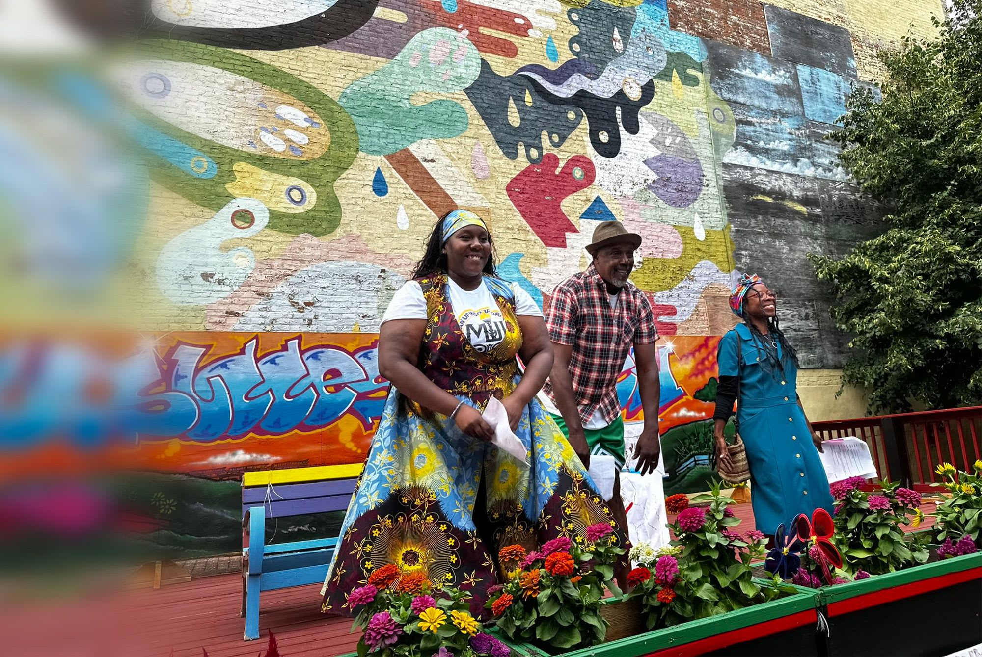 Three performers take a bow during a Roundabout Theatre Company community residency event, surrounded by flowers and a vibrant outdoor mural.