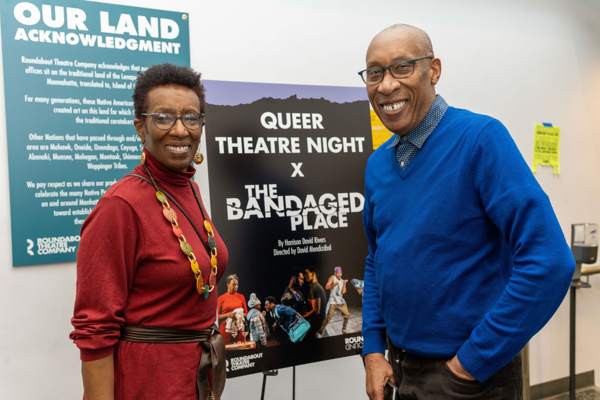 Two guests pose in front of a Queer Theatre Night × The Bandaged Place event poster and a Roundabout Theatre Company land acknowledgment sign.