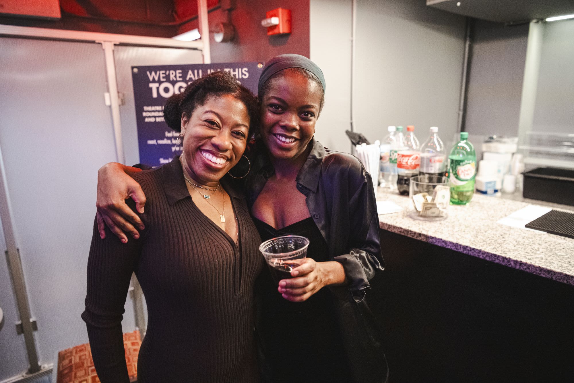 Two guests smile and pose together at Covenant: Black Queer Theatre Night at Roundabout Theatre Company, near a bar area with a "We're All In This Together" sign visible behind them.
