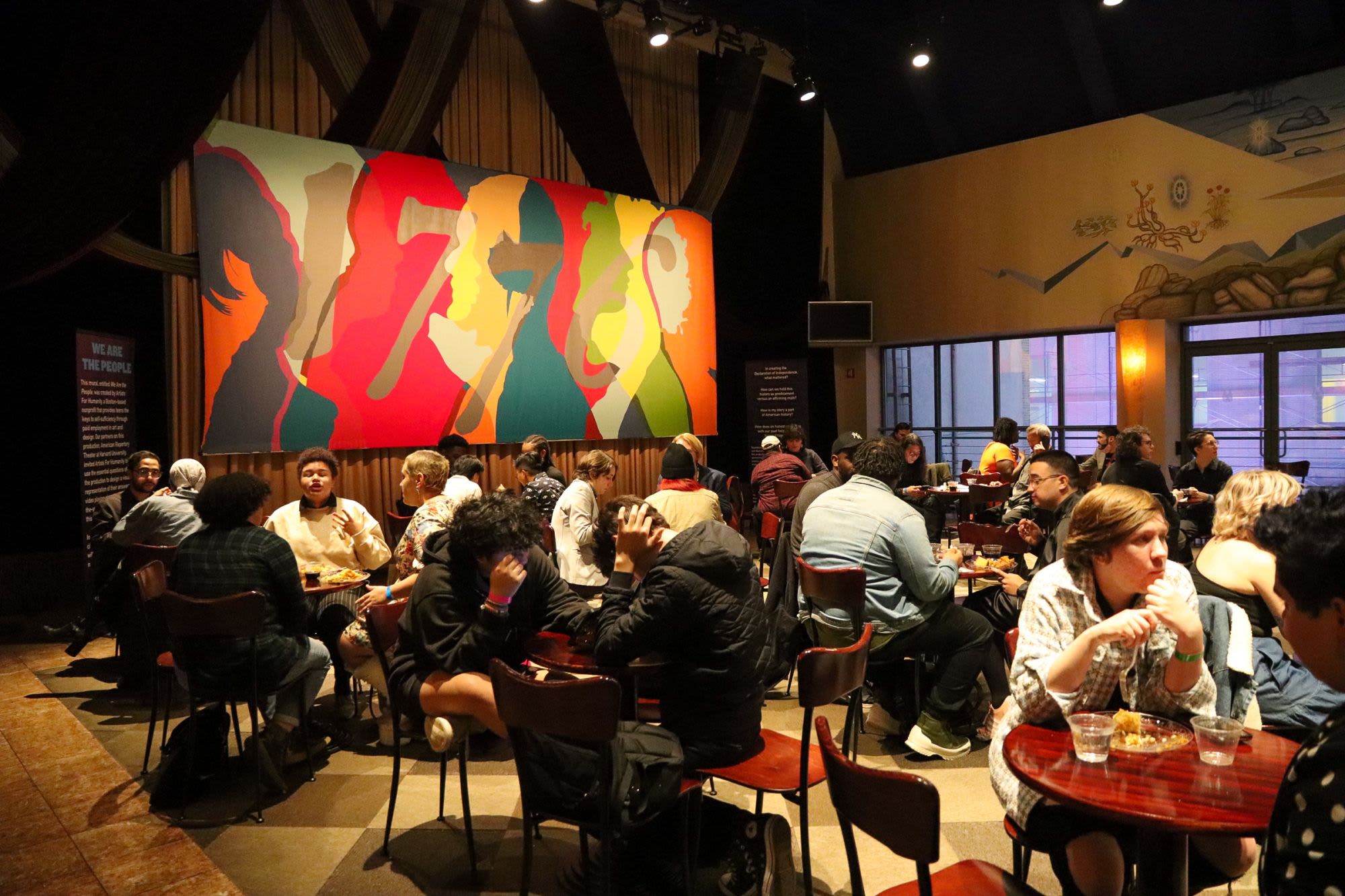 A diverse crowd of guests dines and mingles at a Breaking the Binary Night reception for 1776 at Roundabout Theatre Company, beneath a large colorful mural of silhouetted figures.