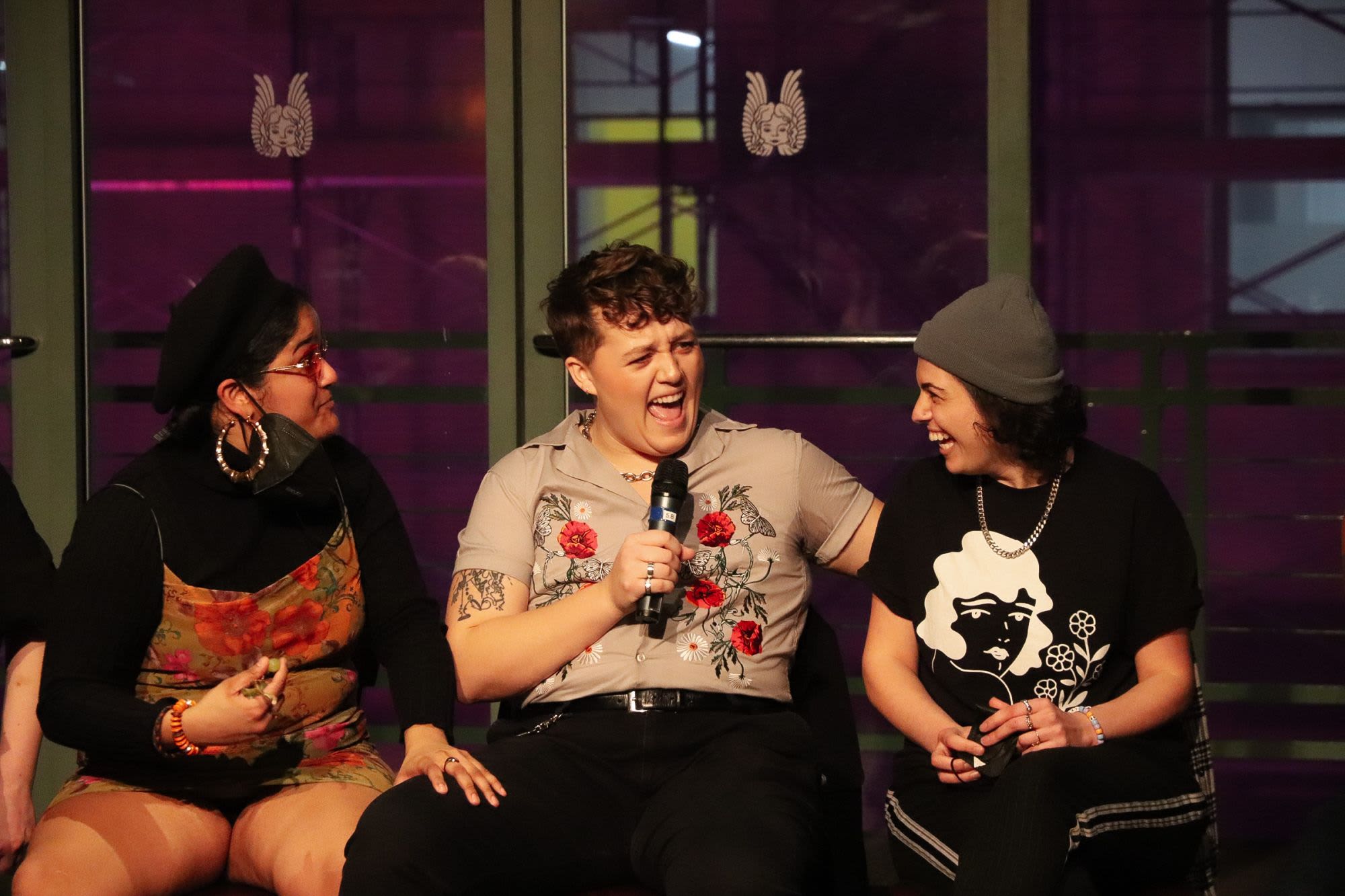 A panelist speaks energetically into a microphone during a Breaking the Binary Night discussion for 1776 at Roundabout Theatre Company, flanked by two other participants.