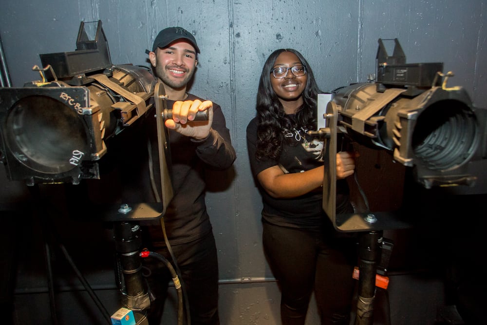 Two students dressed in black stand behind spotlights and smile.