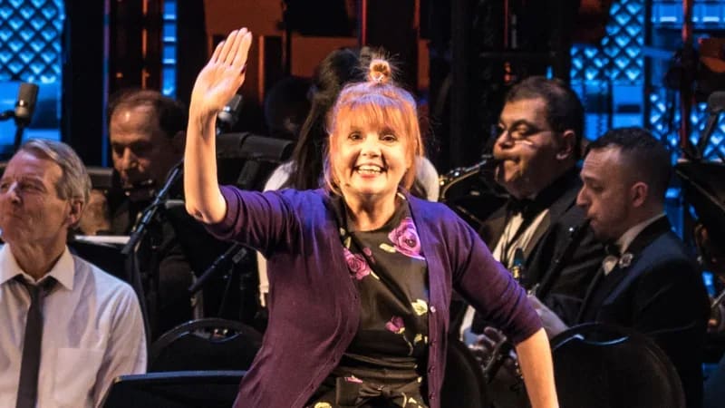 Annie Golden waves on stage in front of an orchestra. She wears a purple cardigan over a black dress with purple roses.