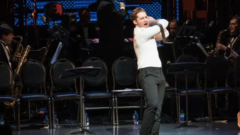 Matthew Morrison pretends to swing a baseball bat. He wears a white collared shirt and black pants.