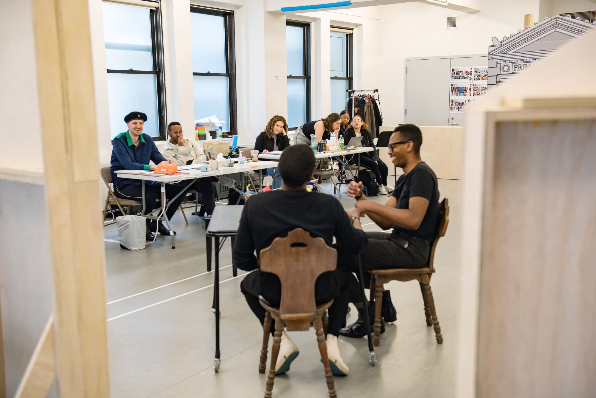 A group of actors perform a scene in a rehearsal studio, the creative team watching in the background.