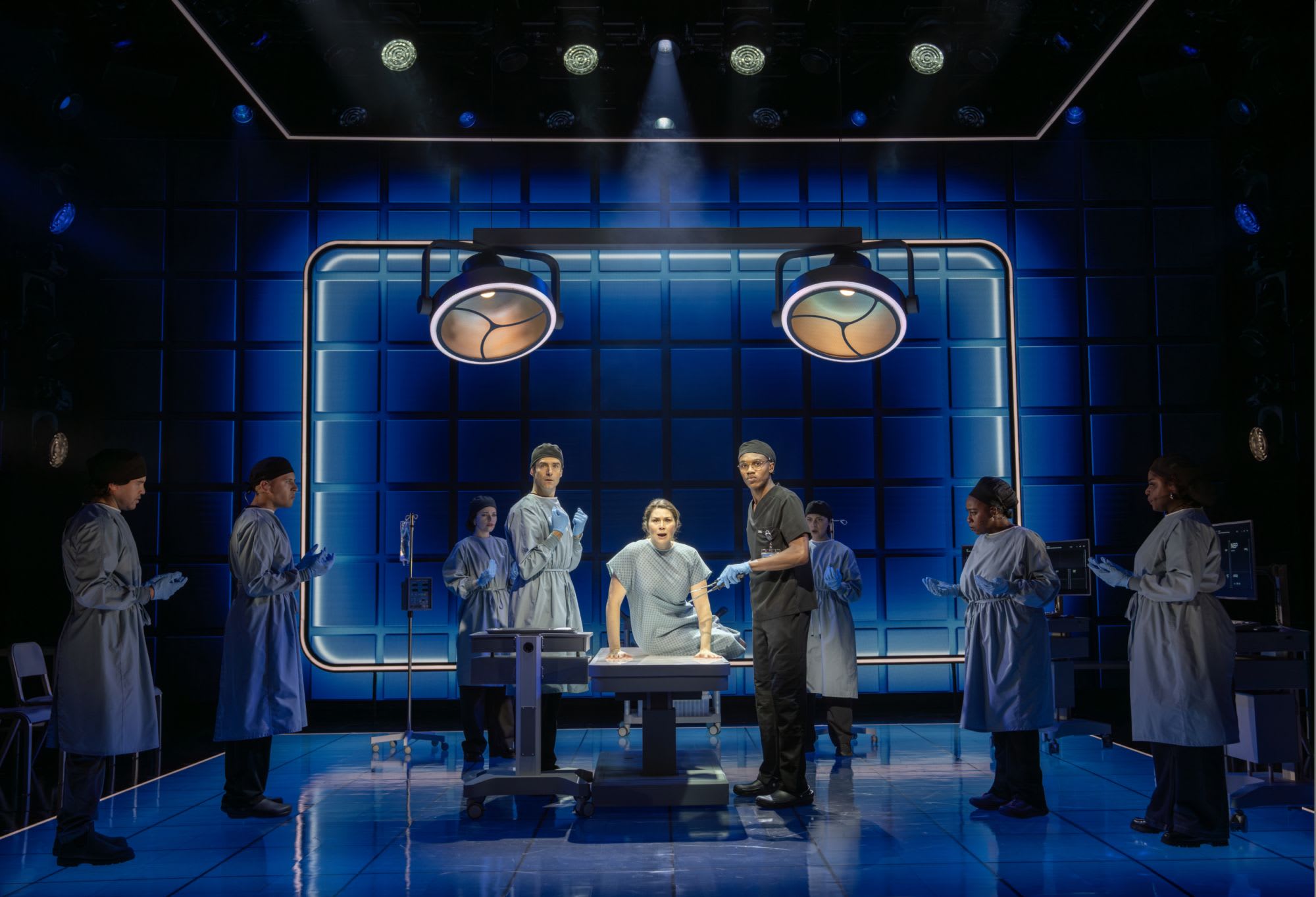 Three actors on stage in surgical gowns in an operation room.