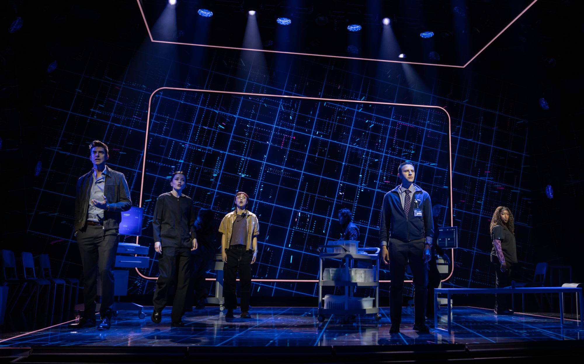 Actors on stage in front of a futuristic video screen with grids.