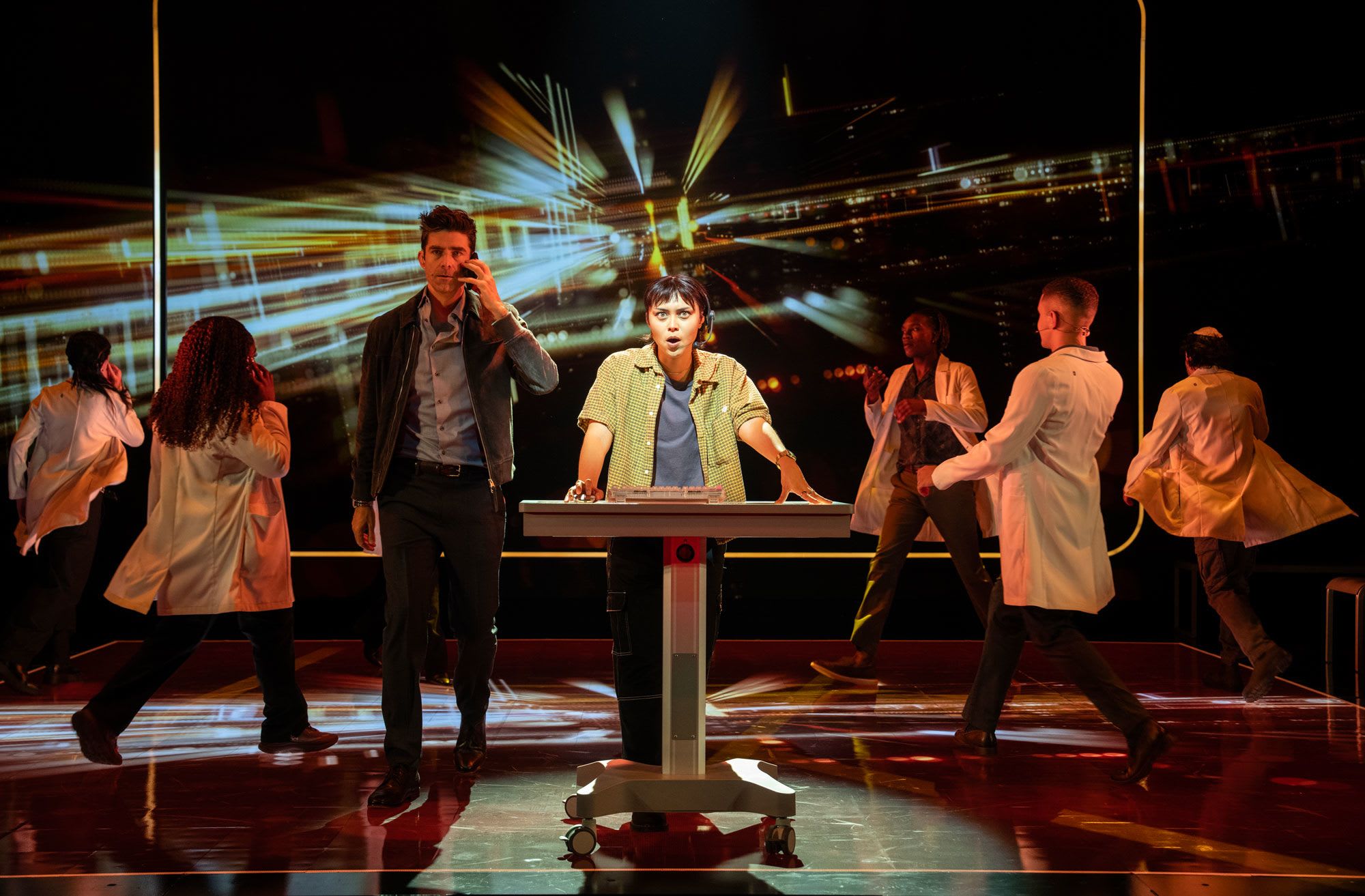 A group of actors onstage, some wearing lab coats, in front of a futuristic video screen.