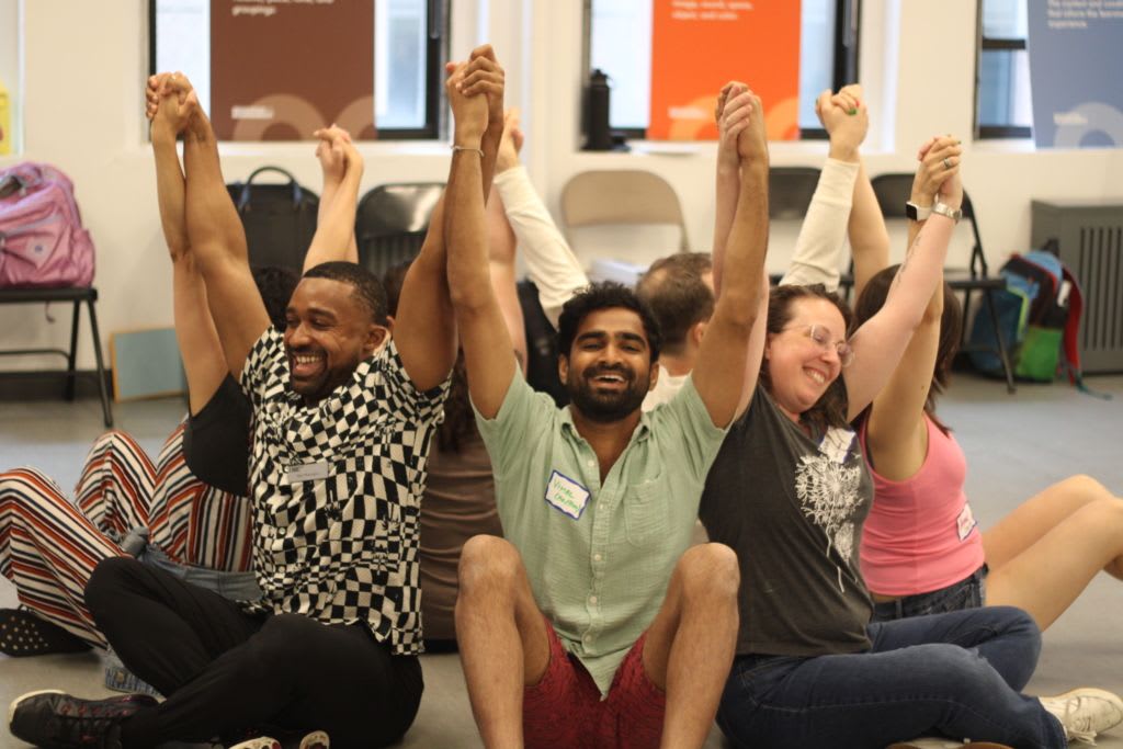 A group of teachers at a workshop sit on the ground facing away from each other in a circle as they hold hands and raise them high in the air.