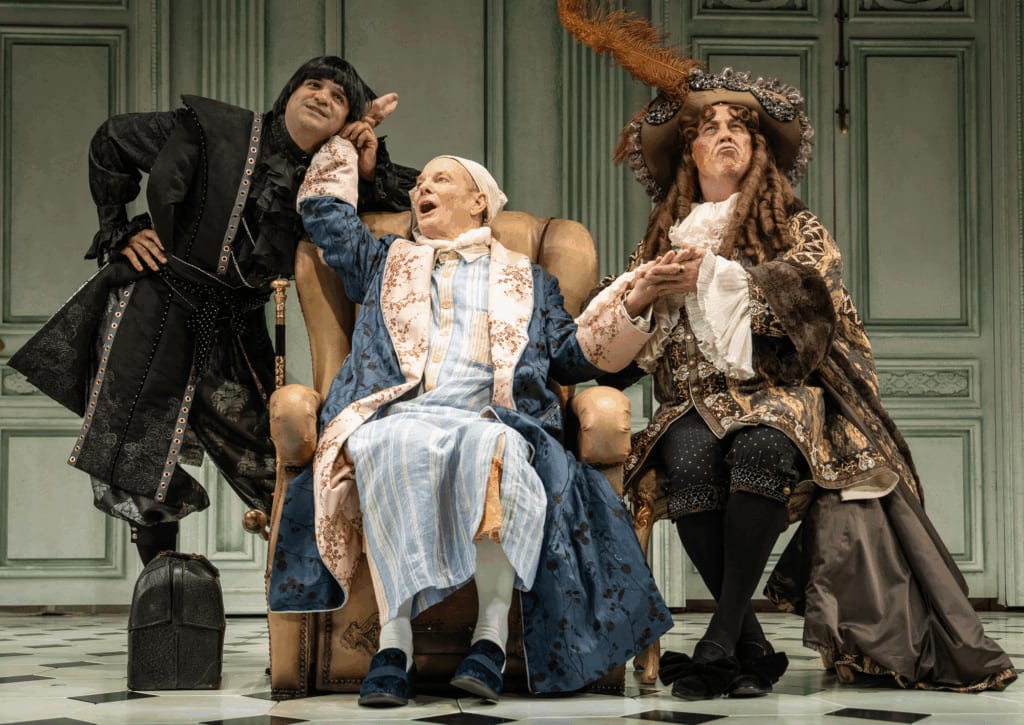Three actors in colorful period piece costumes on an ornate stage. Two are sitting.