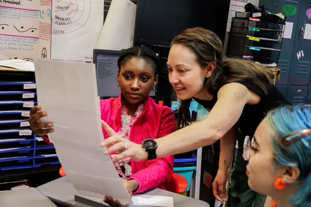 A teaching artist leans over at a students desk to examine an assignment together.