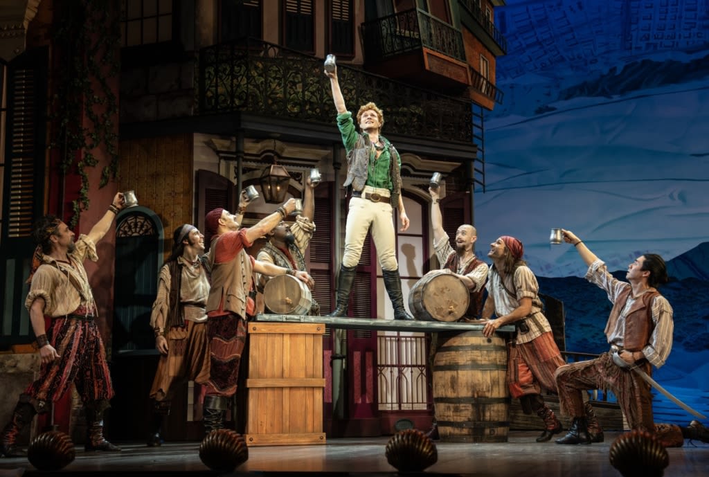 In a New Orleans street setting, a group of men dressed as pirates raise silver mugs in a toast to a young man who stands on a plank balanced on two barrels.