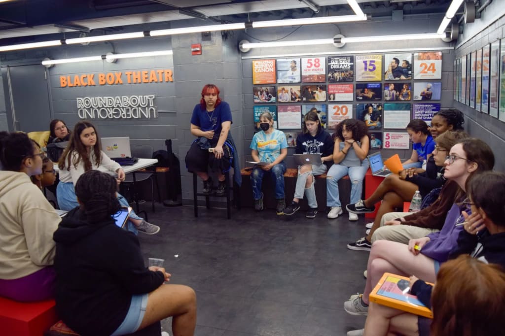 A large group of young people of different skin tones sits in a circle in the lobby of a black box theatre.