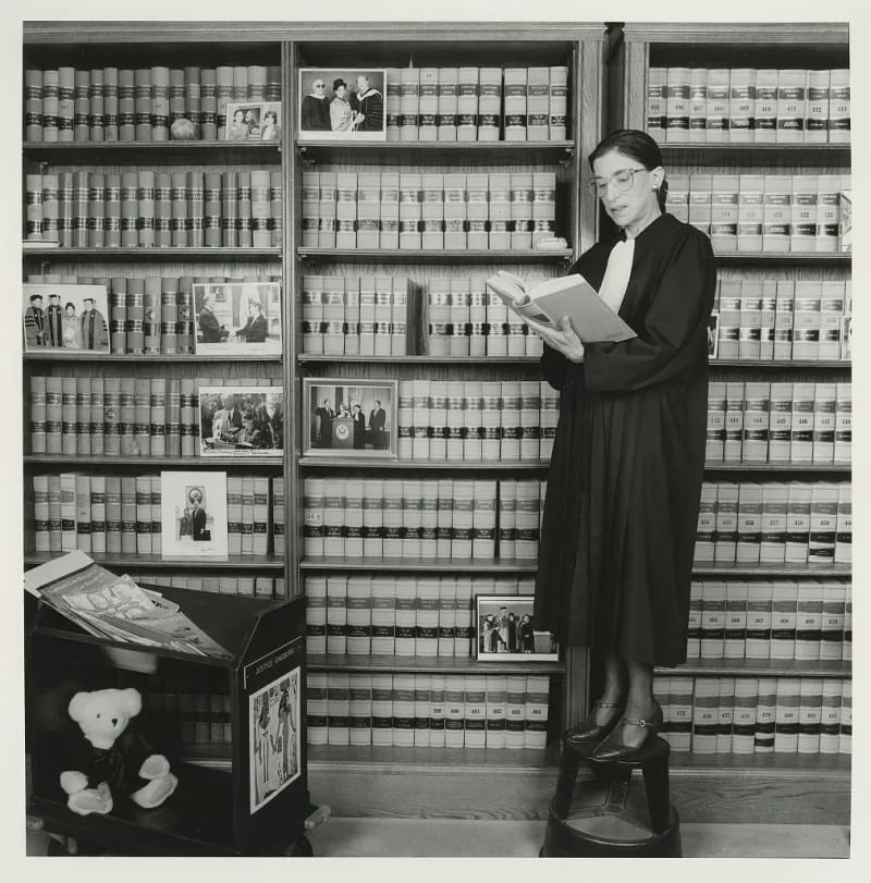 A short woman in a black justice's robe and in glasses stands on a stool reading a book in front of several bookshelves.