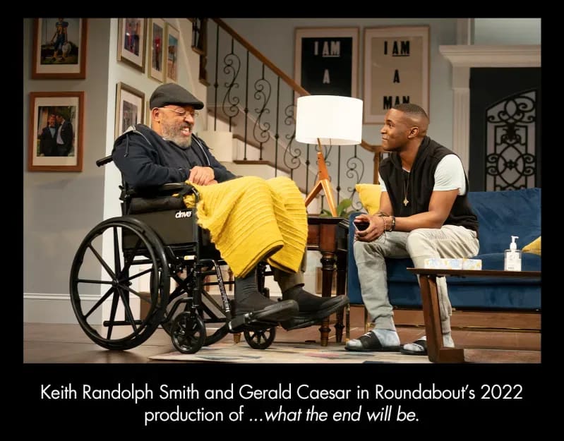 Two men sitting in a living room. There are stairs in the back, and photos on the walls. The man on the left sits in a wheelchair and has a yellow blanket on his lap. The man on the right sits on a blue couch.