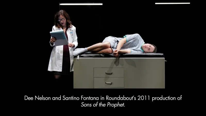 A light-skinned woman in a doctor's coat reads a medical chart while a light-skinned man with dark hair lies on a table in a hospital gown.