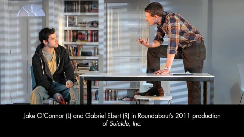 A young, light-skinned man in jeans and a hoodie sits and speaks with a tall, light-skinned man in a plaid button-down shirt. The tall man has his foot on a chair and leans toward the young man.