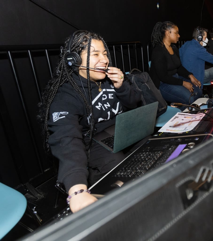 A student in a black shirt smiles while talking into a headset while sitting behind a sound and lighting board.