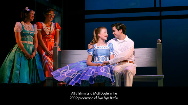 Production photo from BYE BYE BIRDIE. A light-skinned teenage girl wearing a blue dress and crinoline sits on a park bench with a light-skinned, dark-haired teenage boy in a white, collared shirt.