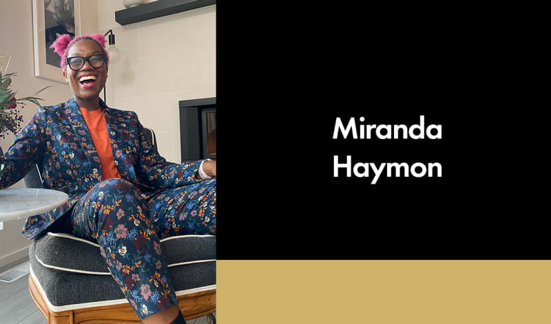 Miranda Haymon, a dark-skinned person with pink hair and glasses smiles. They wear a dark blue suit with colorful flowers over an orange shirt.