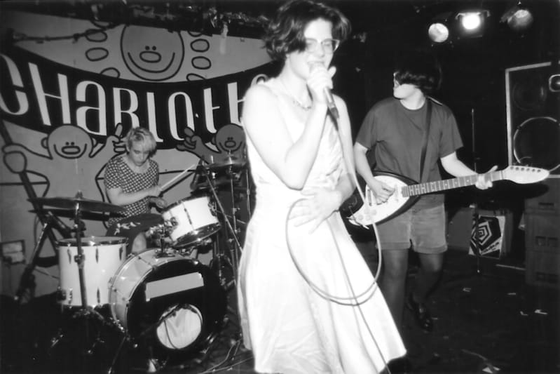 A young, light-skinned woman in a light-colored dress and glasses sings passionately into a wired microphone. Behind, a young woman plays drums and a young man plays guitar.