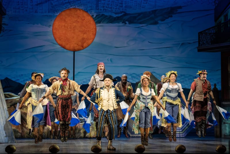 A production image from PIRATES of the song, Modern Major General. An ensemble holds semifore flags behind a man dressed as a general.