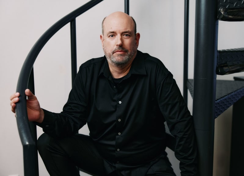A man with light skin and gray facial hair sits on a black spiral staircase, one hand holding the railing. He is partially bald and wears a black button up shirt and black pants.