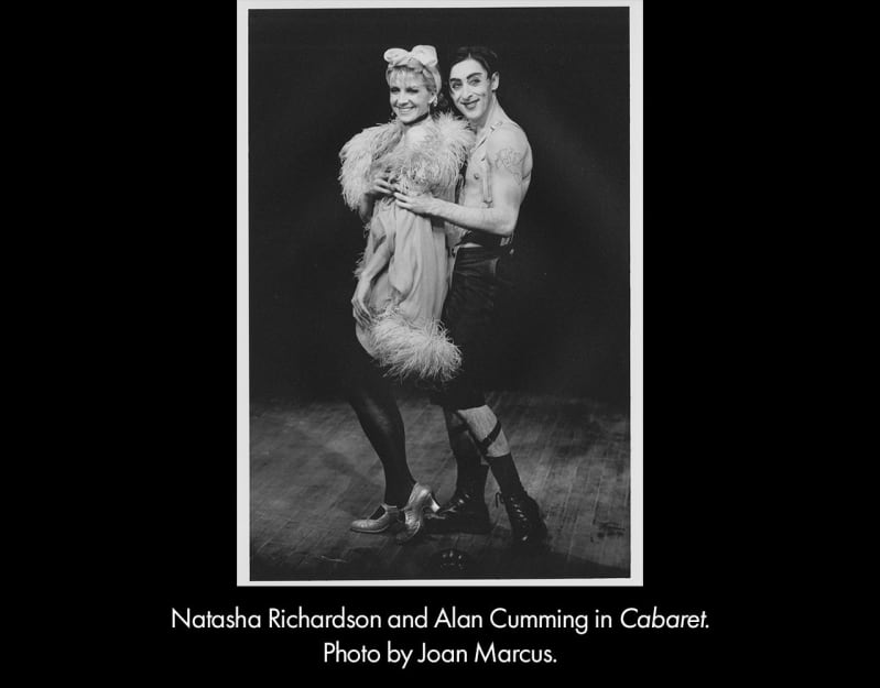 Black and white photo of Natasha Richardson and Alan Cumming in the 1998 Cabaret. She wears a frilly coat and tights and he is shirtless and wears suspenders and black pants.
