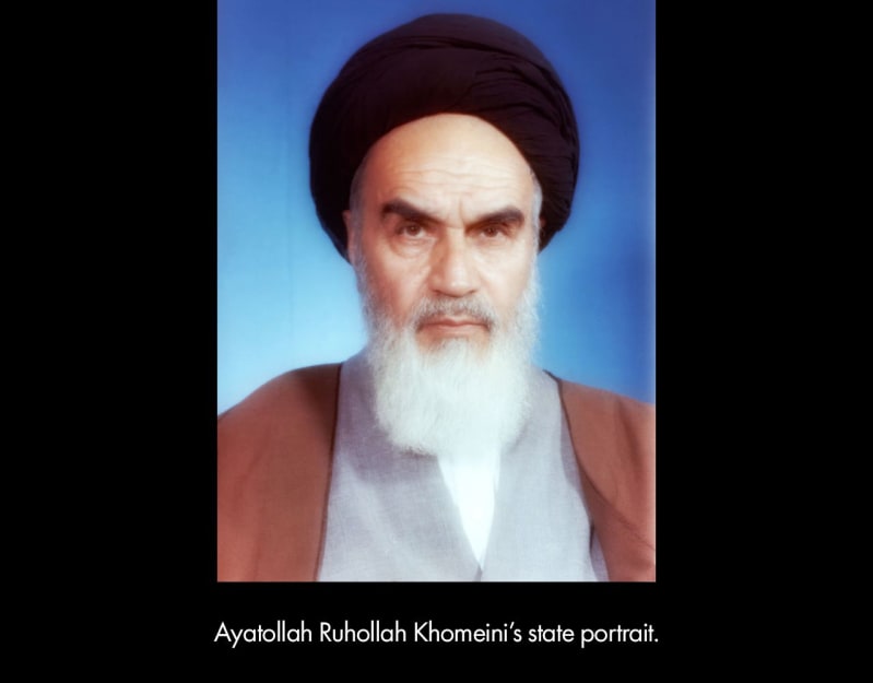 Ayatollah Ruhollah Khomeini's state portrait. An older, light brown-skinned man with a long, white beard wears a black head wrap and poses in front of a blue background.