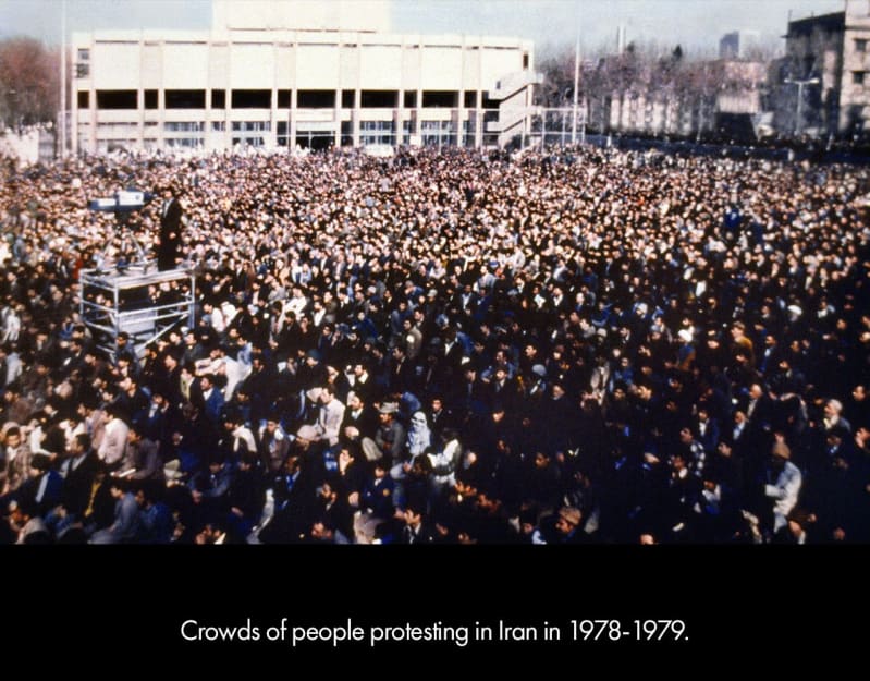 Crowds of people protesting in Iran in 1978-79. Hundreds of people crowd together in front of low-rise buildings.