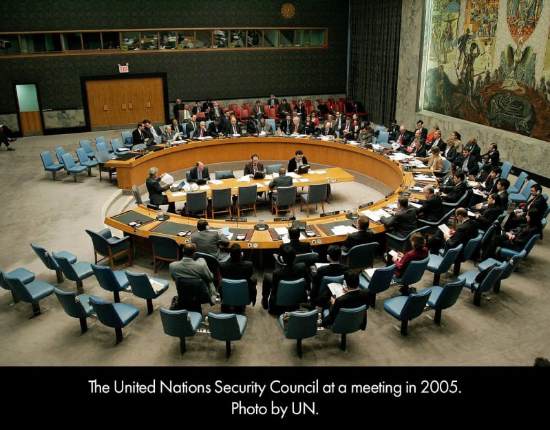 The United Nations Security Council at a meeting in 2005. Formally-dressed adults seated in a large room with three rows of chairs encircling a central table. 