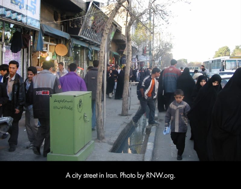 A city street in Iran with a crowded sidewalk bordered on one side by shops and the other by a street full of cars.