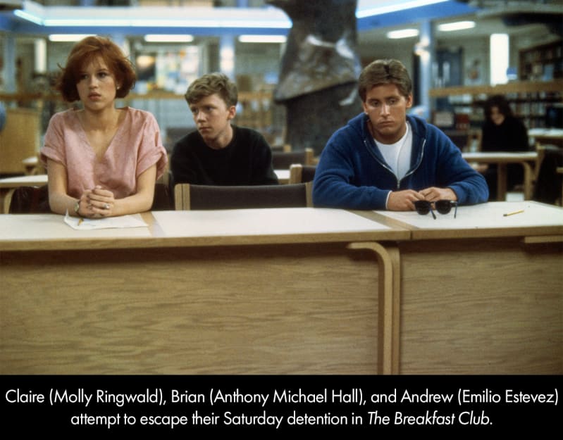 A still from the movie THE BREAKFAST CLUB. A teenage girl with red hair wearing a pink top, a teenage boy in a dark shirt, and a teenage boy in a blue hoodie sit in a school library.
