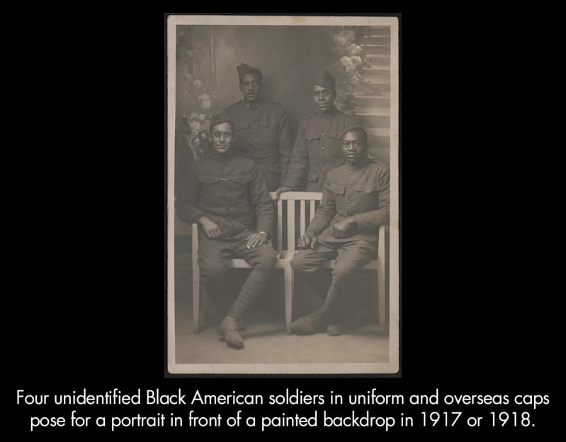 Four unidentified Black American soldiers in uniform and overseas caps pose for a portrait in front of a painted backdrop in 1917 or 1918