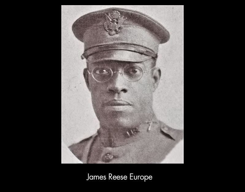 Black and white photograph of James Reese Europe. A dark-skinned man in round, wireframe glasses and a military cap.