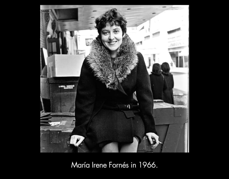 María Irene Fornés in 1966. A light-skinned woman with short, dark hair and dark eyes smiles into the camera. She is seated on top of a large wooden crate. She wears a dark coat with a fur collar. 