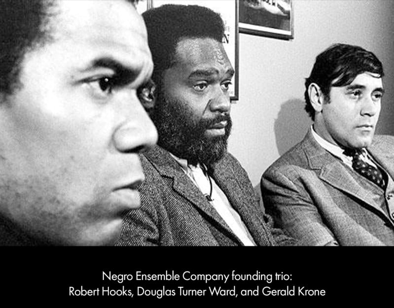 Foreground, the face of a dark-skinned man. To his right is a dark-skinned man with a full beard, wearing a suit, and a light-skinned man with dark hair, wearing a suit.