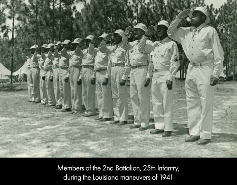 A line of dark-skinned men in light-colored military uniforms and hats. They salute with their right hands.