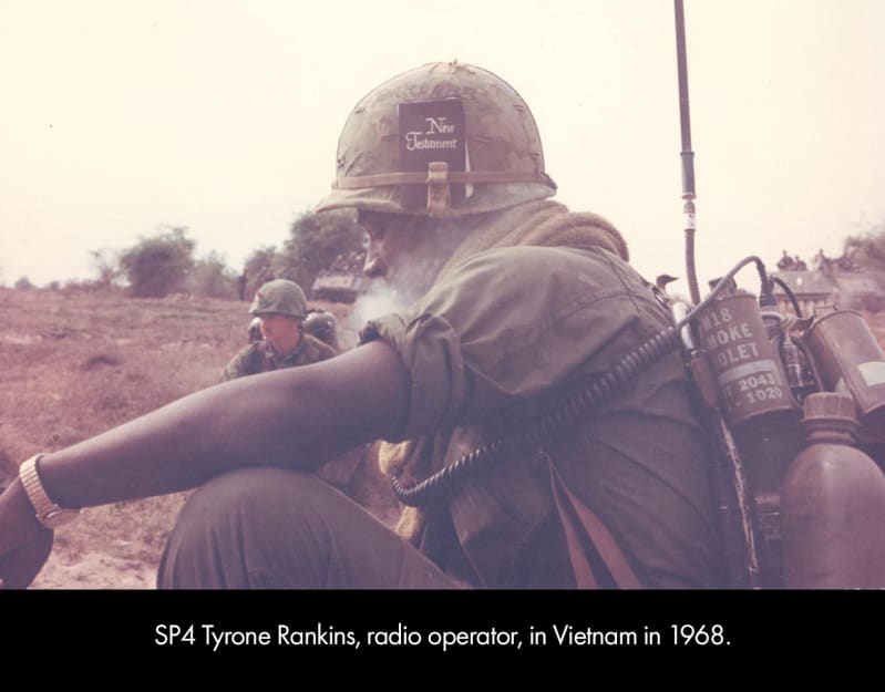 A young, dark-skinned man in a drab green military uniform sits. His helmet has a black book with the words “New Testament” stuck in the brim. He wears a backpack with metal canisters in it, a gold wa
