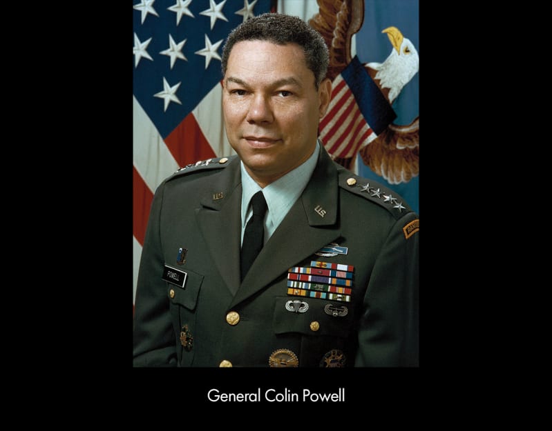 A medium-dark skinned man with graying hair wears a green military dress jacket with four stars across his shoulders and five colorful stripes on the left breast above several medals.