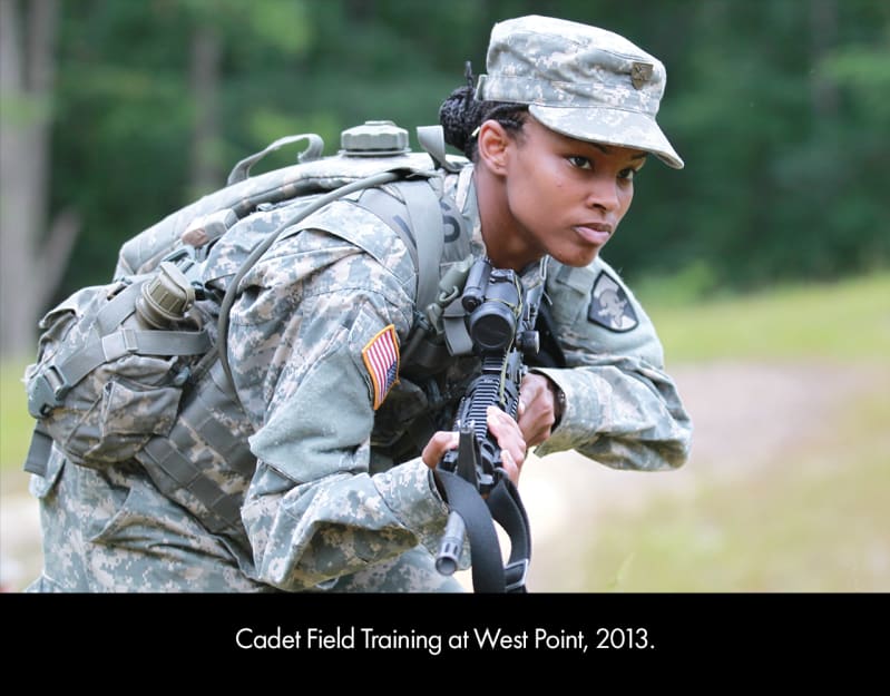 A young, dark-skinned woman in a green camouflage hat, pants, and jacket. There is an American flag on the right arm of the jacket. She holds a black gun and leans forward, watching.