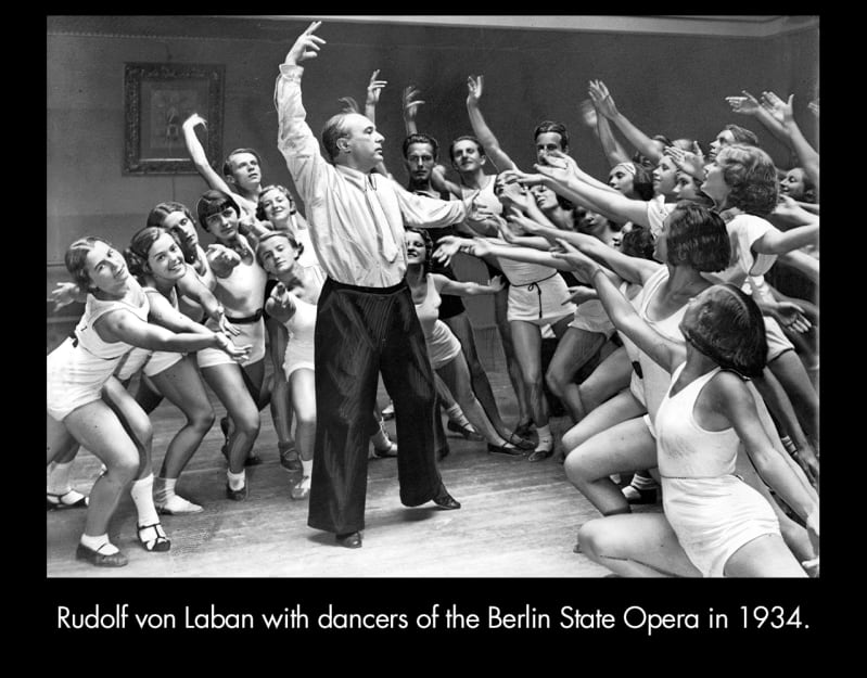  Rudolf von Laban with dancers of the Berlin State Opera in 1934. A light-skinned man in wide-leg pants and a short tie raises his arm above his head. He is surrounded by women in leotards.