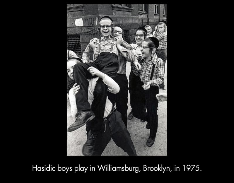Hasidic boys play in Williamsburg, Brooklyn, in 1975. A group of light-skinned boys with long sidelocks, wearing yarmulkes and long-sleeved oxford shirts, laugh as they lift one boy into the air. 