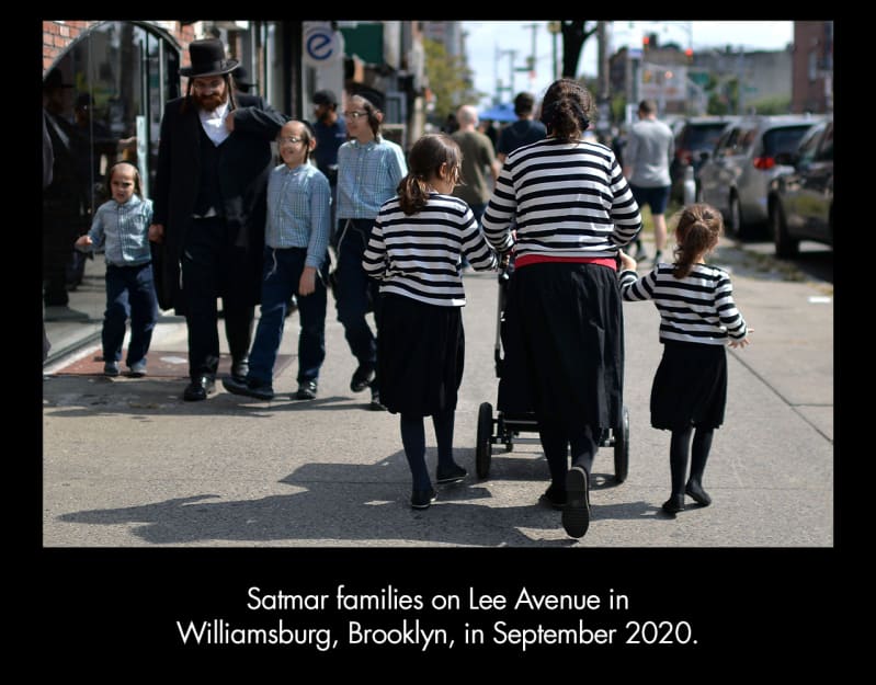 Satmar families on Lee Avenue in Williamsburg, Brooklyn, in September 2020. A light-skinned man with long sidelocks and a beard, wearing a dark suit and hat, walks with three young light-skinned boys 