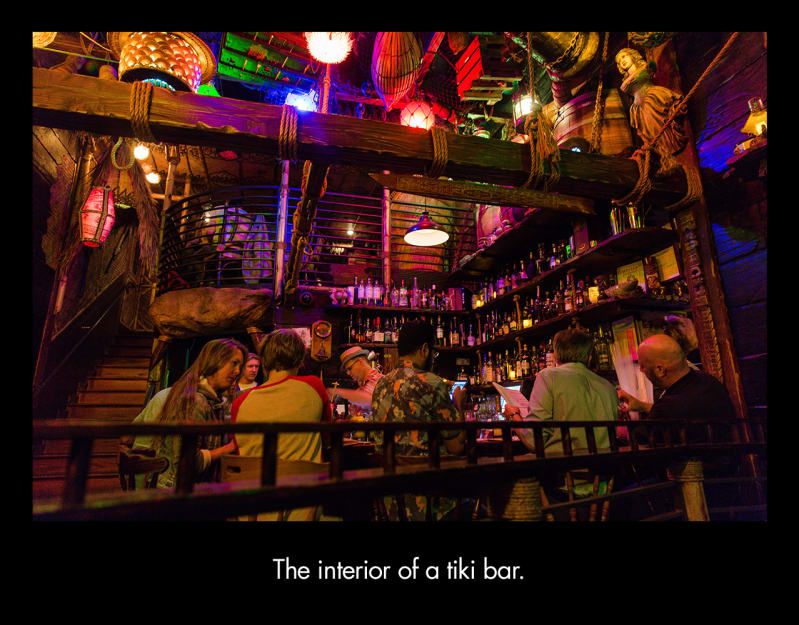 Six adults sit at a wooden bar. Bottles line the walls behind them. Above the bar is a rough wooden beam with ropes around it and colorful lights.  