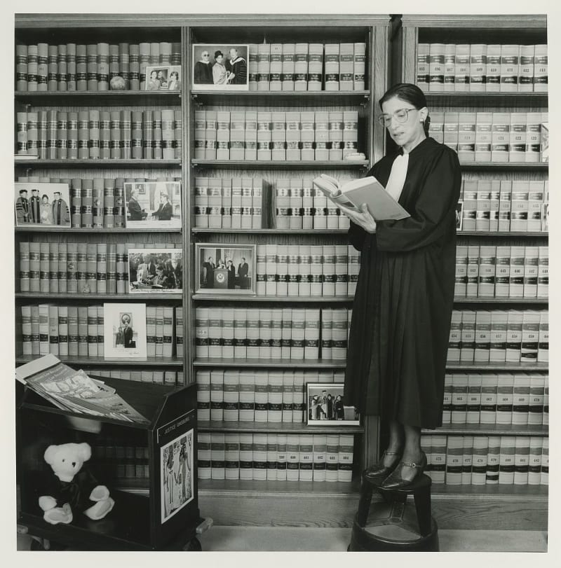 A petite light-skinned woman in a black robe and glasses stands on a stool in front of several bookcases. 