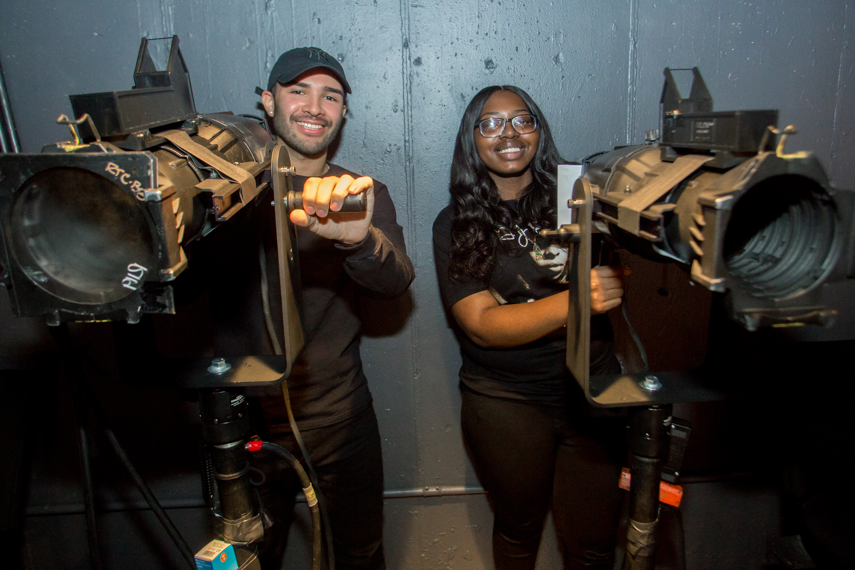 Two students dressed in black stand behind spotlights and smile.