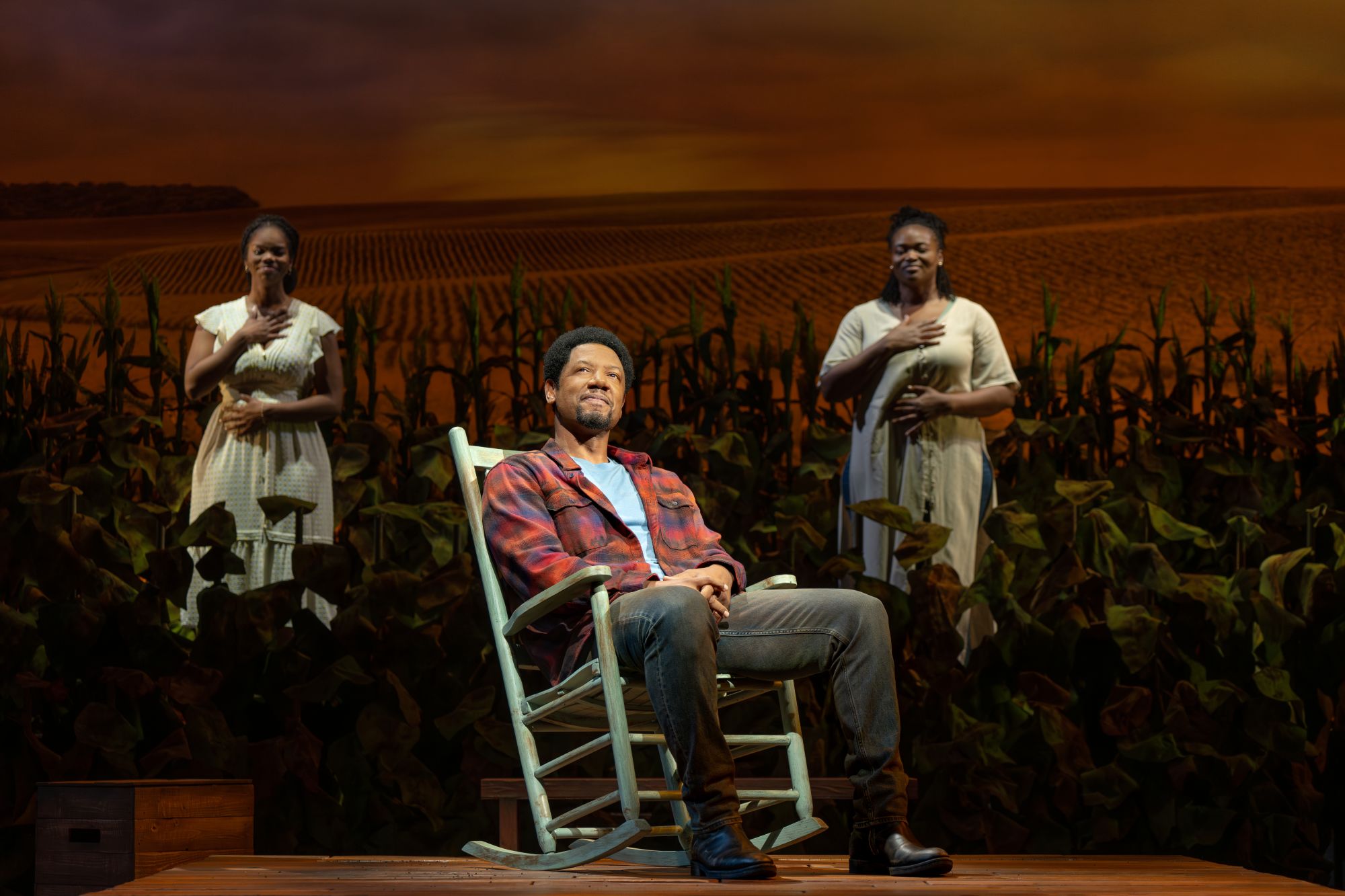 A production photo from HOME. A man sits in a rocking chair while two women stand in a field of crops behind him with hands over their hearts.
