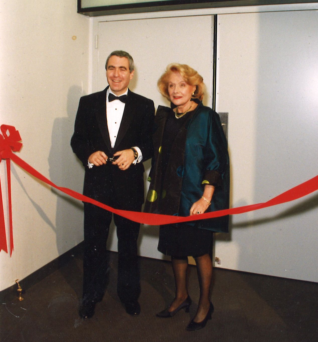 Todd Haimes in a tuxedo (left) and Laura Pels in a dark dress (right) cutting a red ribbon together in front of the white doors of the Laura Pels Theatre.